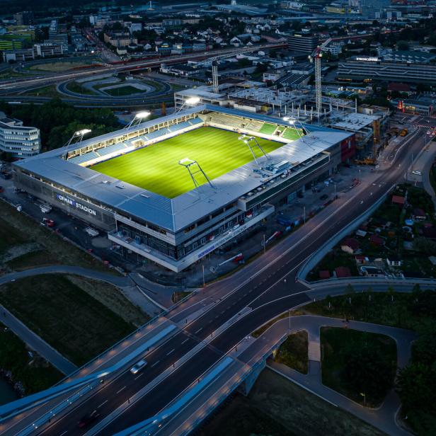 Ein beleuchtetes Fußballstadion mit grünem Spielfeld liegt an einer großen Straßenkreuzung in einer Stadt bei Nacht.