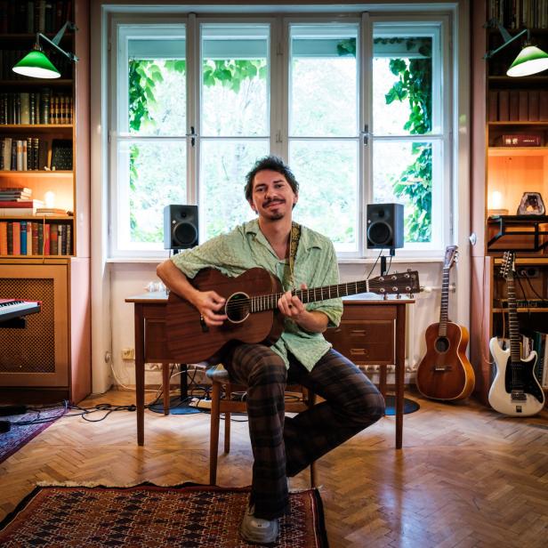 Ein Mann mit Gitarre sitzt in einem Raum voller Bücher und Musikinstrumente.