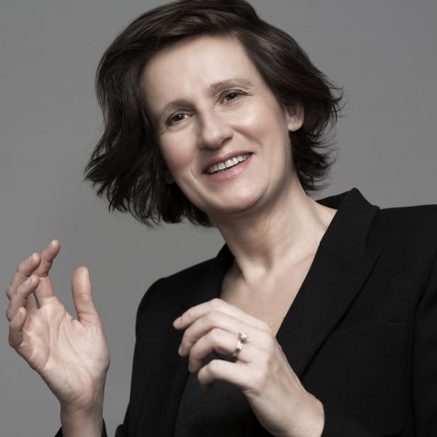 Anna Flipo lächelt in einem schwarzen Blazer vor einem grauen Hintergrund.
