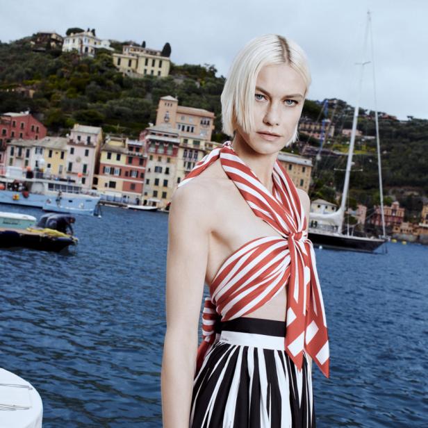 Eine Frau mit blonden Haaren, gekleidet in ein gestreiftes Outfit, vor der Küstenstadt Portofino.