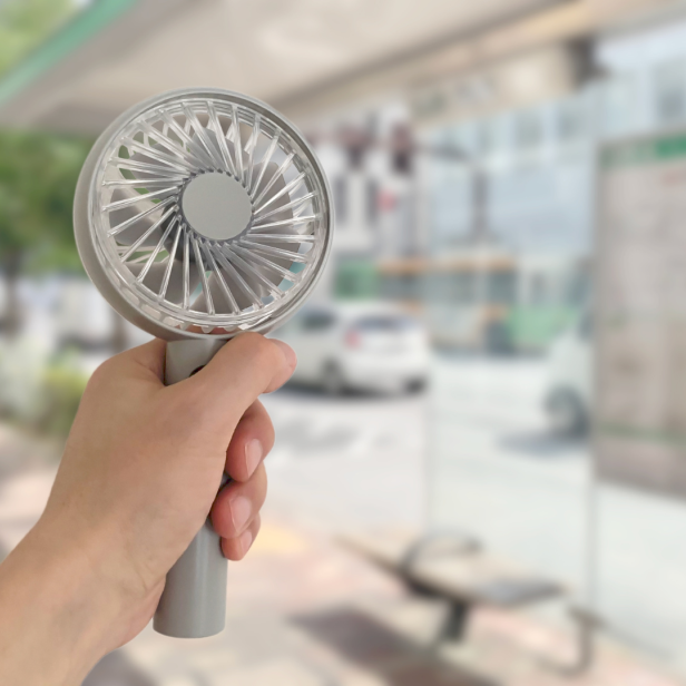 Eine Hand hält einen kleinen, grauen Ventilator an einer Bushaltestelle.