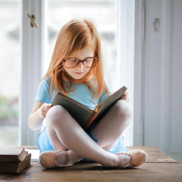 Ein rothaariges Mädchen mit Brille liest ein Buch auf einem Tisch.