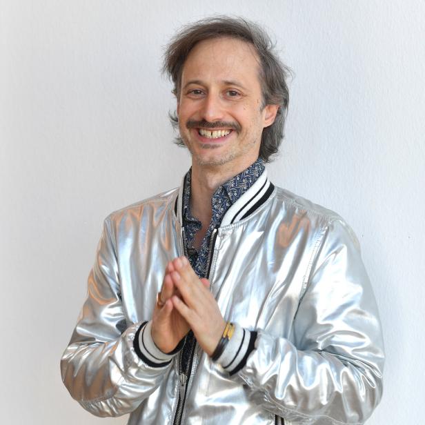 Ein lächelnder Mann mit Schnurrbart und silberner Jacke steht vor einer weißen Wand.