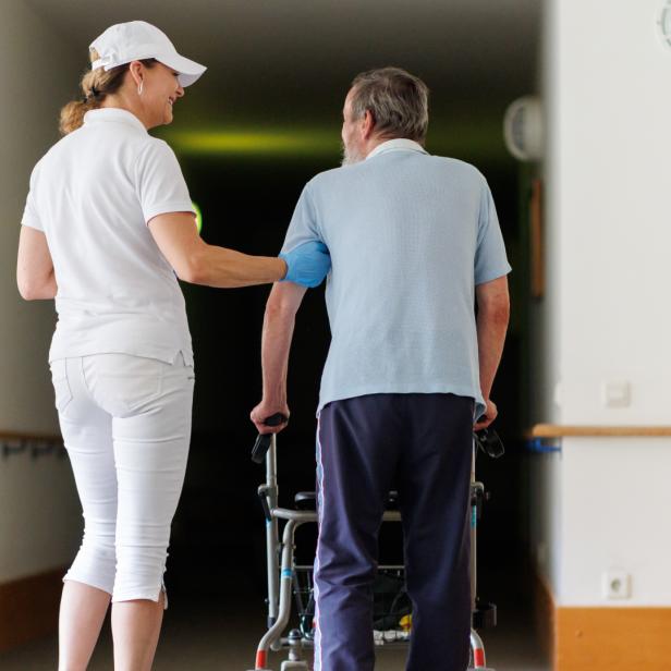 Eine Pflegerin unterstützt einen älteren Mann mit Rollator in einem hellen Flur.