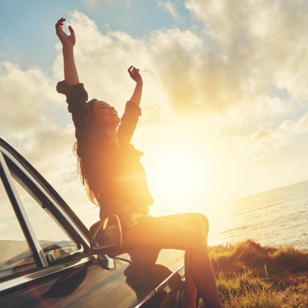 Eine junge Frau sitzt auf einem Auto am Meer und streckt die Arme in die Luft.
