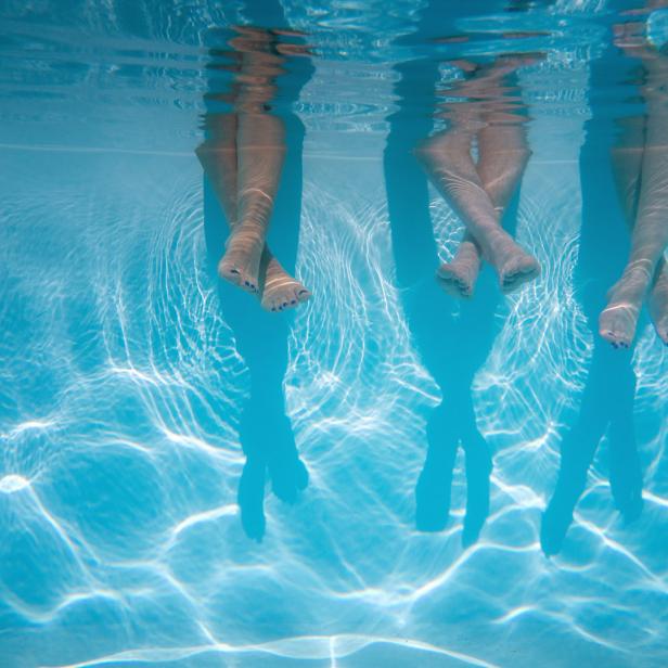 Drei Paar Beine ragen ins blaue Wasser eines Schwimmbeckens.