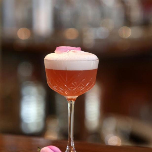 Ein rosa Cocktail mit Schaum und Rosenblättern auf einer Bar.