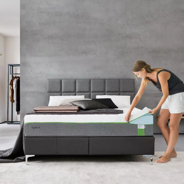 Eine Frau bezieht ein modernes Bett mit grauem Kopfteil in einem minimalistisch eingerichteten Schlafzimmer.