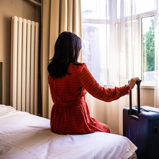 Eine Frau sitzt mit einem Koffer auf einem Hotelbett und schaut aus dem Fenster.
