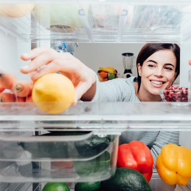 Eine Frau greift lächelnd nach einer Zitrone in einem Kühlschrank, der mit Gemüse und Obst gefüllt ist.