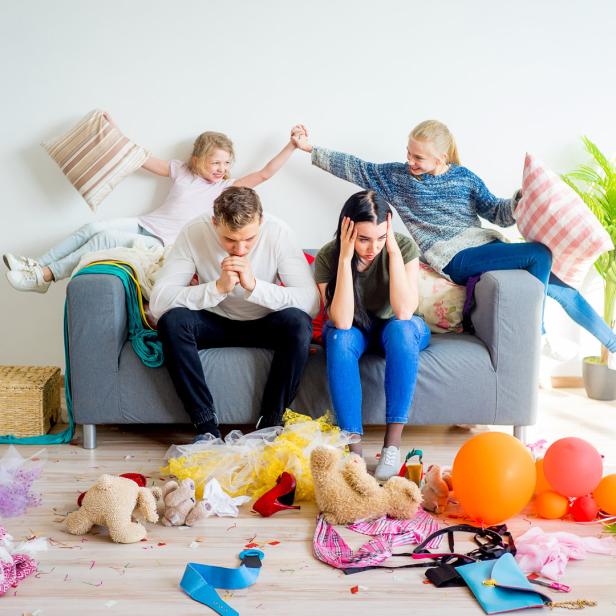 Ein Paar sitzt gestresst auf einem Sofa, während Kinder im Chaos spielen.