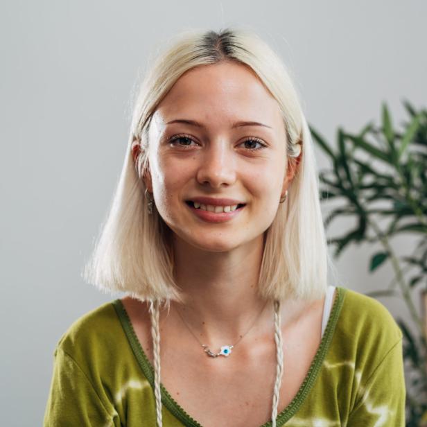 Eine lächelnde junge Frau mit blonden Haaren und einem grünen Oberteil vor einem hellen Hintergrund.