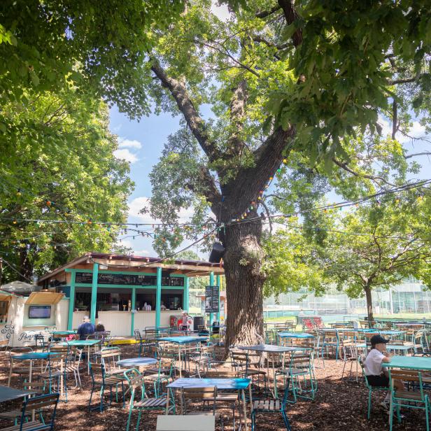 Ein Biergarten mit Tischen und Stühlen unter schattenspendenden Bäumen.