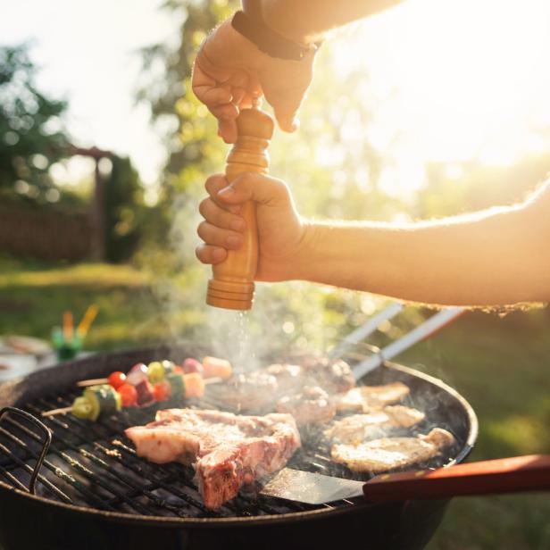 Eine Person würzt Fleisch und Gemüsespieße auf einem Grill.