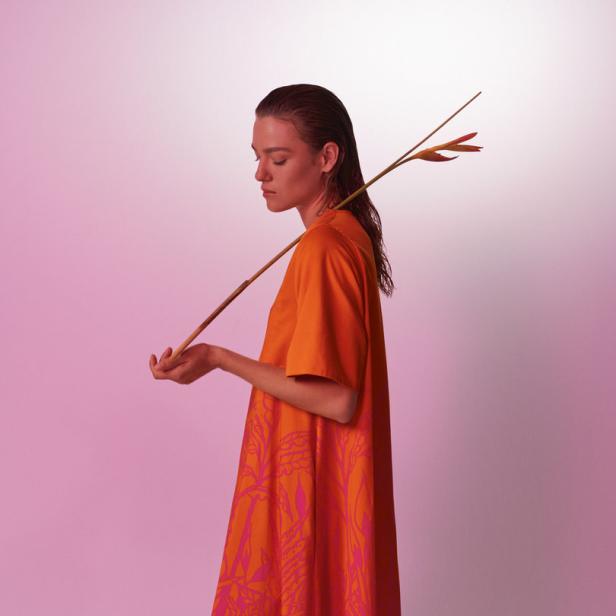 Eine Frau in einem orange-pinkfarbenen Kleid hält eine Helikonienblüte vor einem rosa Hintergrund.
