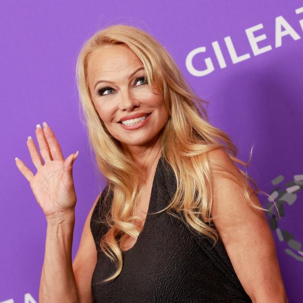 Pamela Anderson winkt vor einem lila Hintergrund.