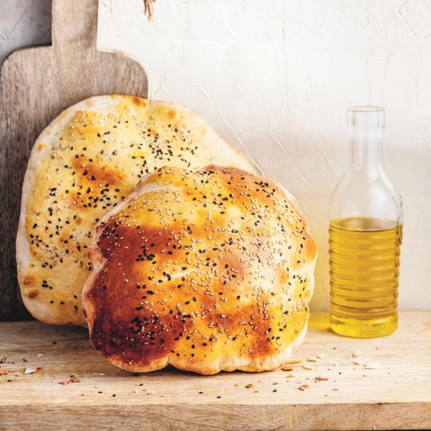 Zwei Pita-Brote mit Sesam und Schwarzkümmel auf einem Holzbrett mit einer Flasche Öl.