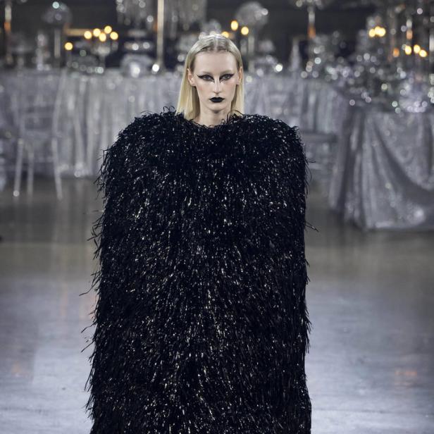 Ein Model mit dunklem Make-up trägt ein schimmerndes, schwarzes Kleid auf dem Laufsteg.