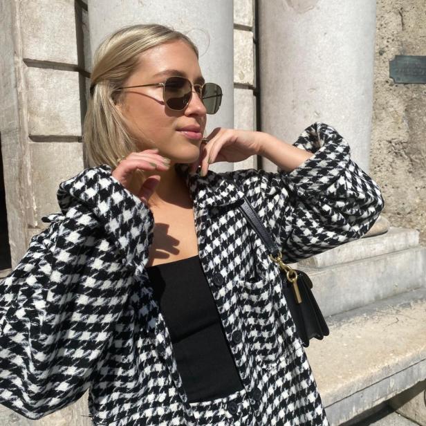 Eine Frau mit Sonnenbrille und Hahnentrittmuster-Jacke posiert vor einem Gebäude.