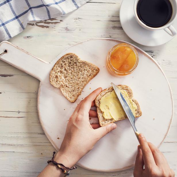 Eine Person bestreicht ein Toastbrot mit Butter, dazu Kaffee und Marmelade.