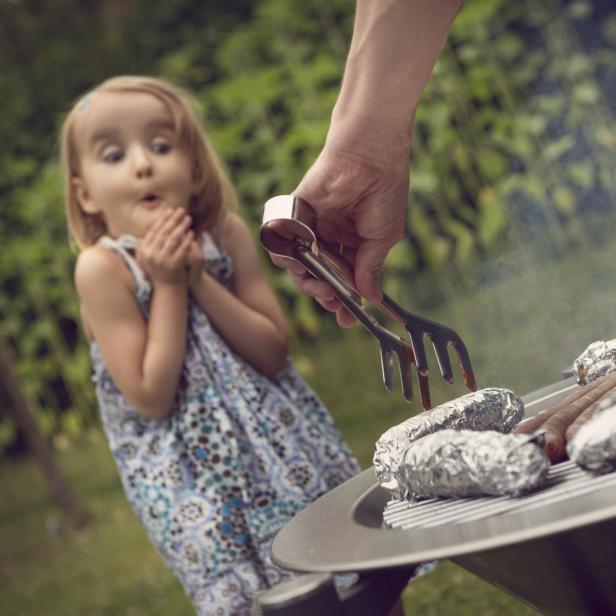 Ein Mann grillt Würstchen und in Folie gewickelte Speisen, während ein kleines Mädchen überrascht zusieht.