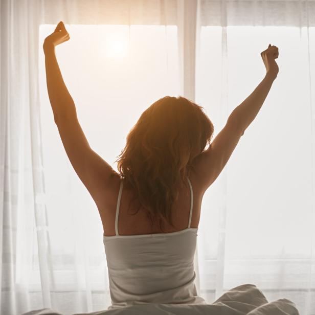 Eine Frau streckt sich morgens im Bett vor einem hellen Fenster.