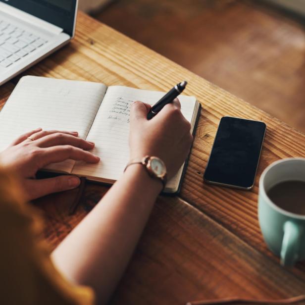Eine Person schreibt in ein Notizbuch auf einem Holztisch mit Laptop, Handy und Kaffeetasse.