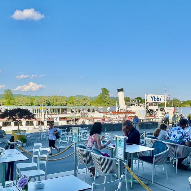 Ein Ausflugsdampfer der Marke Ybbs liegt vor einem Restaurant mit Gästen an Tischen.