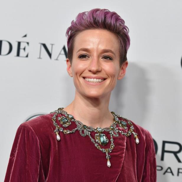 Megan Rapinoe lächelt in einem bordeauxroten Kleid mit Schmuck am Kragen.