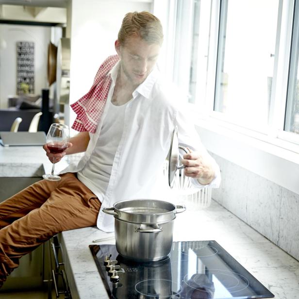 Ein Mann sitzt auf der Küchentheke, hält ein Glas Wein und kocht Pasta.