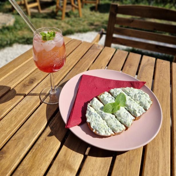 Ein Aperol Spritz und ein Gurkenbrot auf einem Holztisch im Freien.