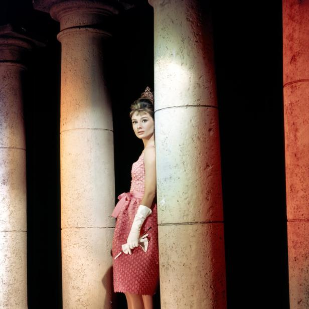 Audrey Hepburn in einem rosa Kleid, posierend zwischen Säulen.