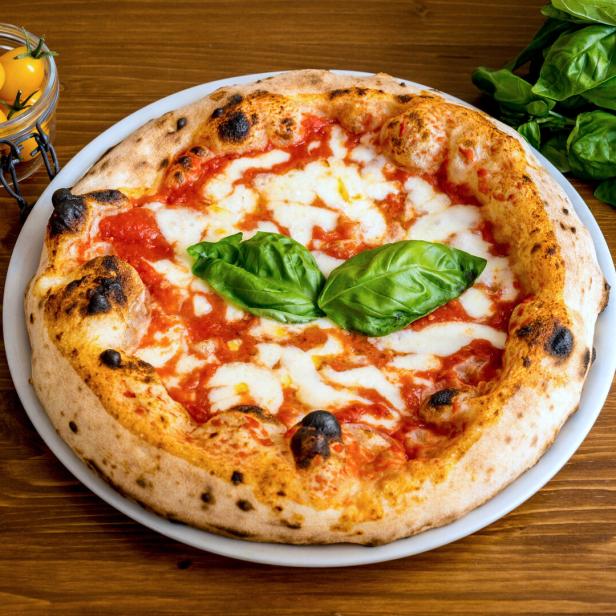 Eine Pizza Margherita mit Basilikum auf einem Teller, dazu Tomaten in Gläsern.