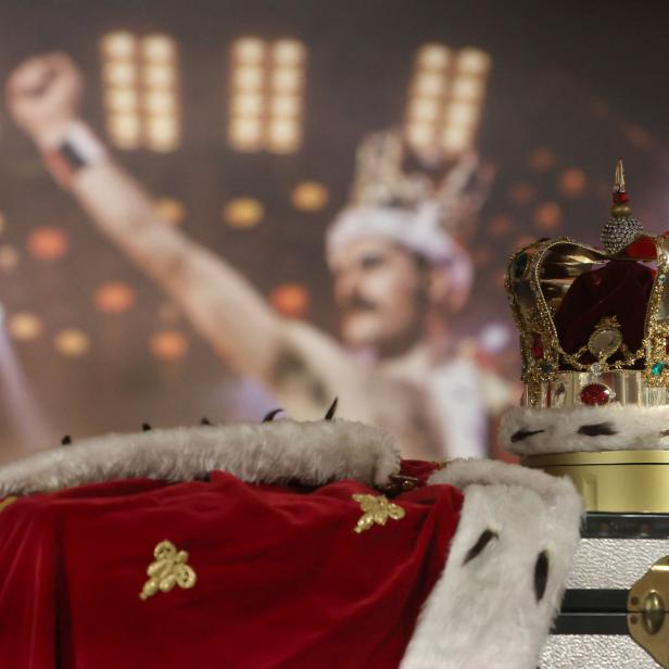 Eine Krone und ein roter Umhang liegen vor einem verschwommenen Bild von Freddie Mercury.