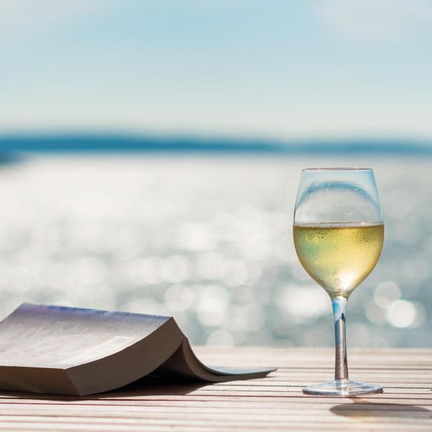 Ein Glas Wein und ein Buch auf einem Holztisch mit Blick auf das Meer.