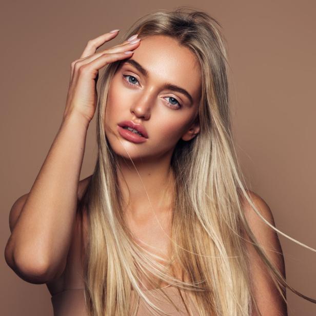 Eine blonde Frau mit blauen Augen posiert vor einem beigen Hintergrund.