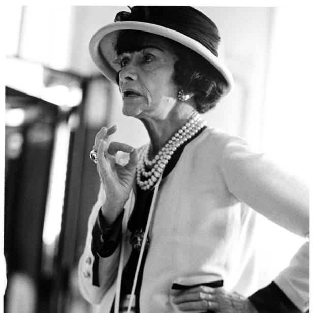 Coco Chanel mit Hut, Perlenkette und Blazer in einer Schwarzweißaufnahme.