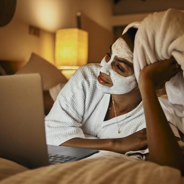 Eine Frau entspannt mit Gesichtsmaske und Laptop im Bett.