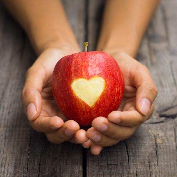 Hände halten einen roten Apfel mit einem ausgeschnittenen Herz auf einem Holztisch.