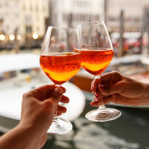 Zwei Personen stoßen mit Aperol Spritz vor einer venezianischen Wasserstraße an.