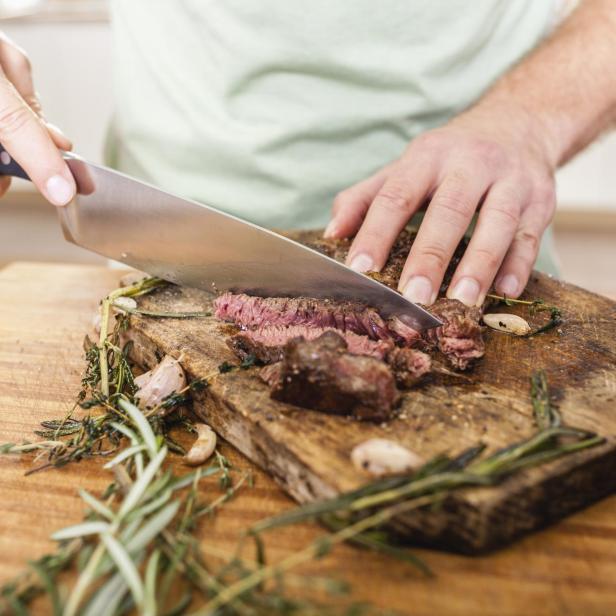 Ein Mann schneidet ein Steak auf einem Holzbrett mit Kräutern und Knoblauch.