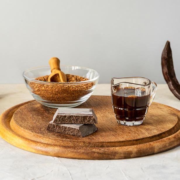 Johannisbrotmelasse, Mehl, Schokolade und Johannisbrotschoten auf Holzschneidebrett mit hellgrauem Hintergrund und Kopierraum - Stock-Fotografie.
