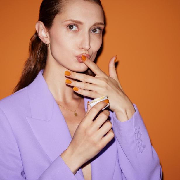 Eine Frau im lila Blazer probiert eine orangefarbene Creme.