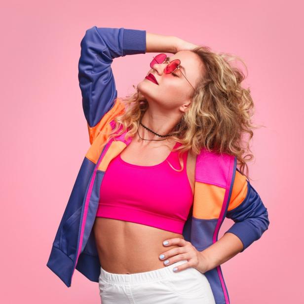 Eine Frau mit Sonnenbrille posiert in Sportkleidung vor einem rosa Hintergrund.