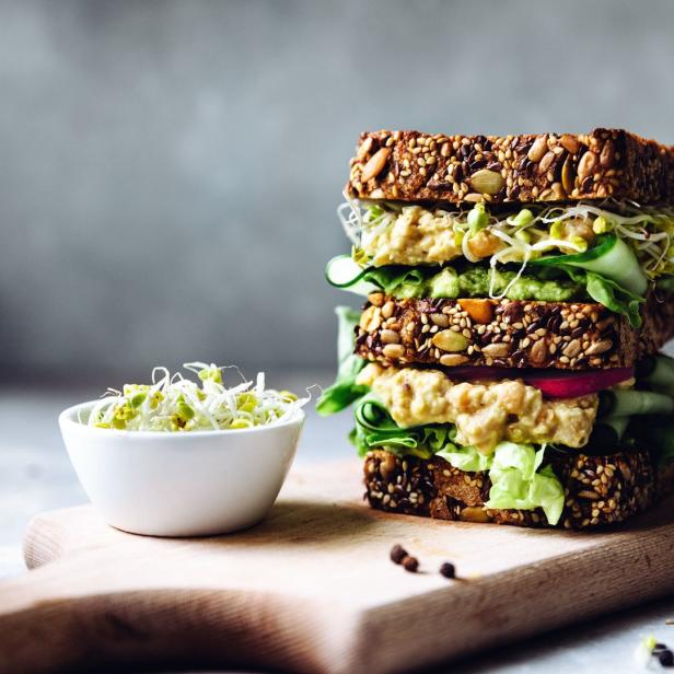 Ein gestapeltes Sandwich mit Hummus, Avocado, Gurke und Sprossen auf einem Holzbrett.