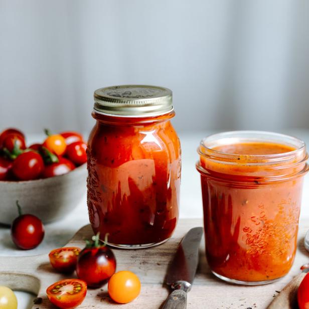 Zwei Gläser Tomatensauce stehen auf einem Schneidebrett mit bunten Tomaten und einem Messer.