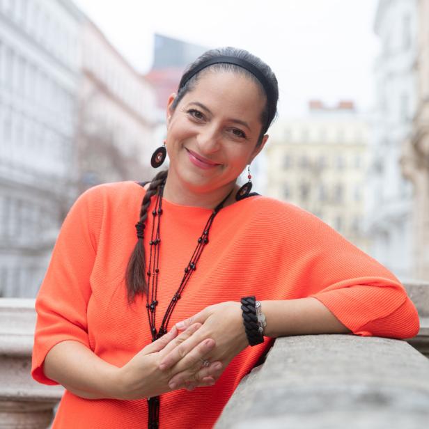 Eine lächelnde Frau in einem orangefarbenen Pullover lehnt an einer Balustrade.