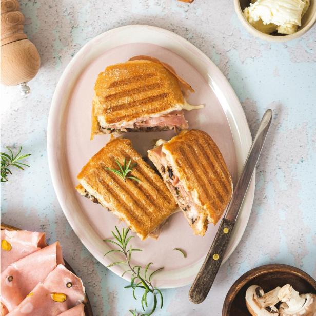 Ein angeschnittenes Panini mit Mortadella und Pilzen auf einem Teller angerichtet.