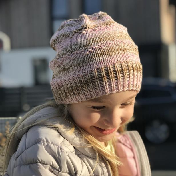 Ein lächelndes Mädchen trägt eine rosa-braun gestreifte Mütze und eine graue Jacke.