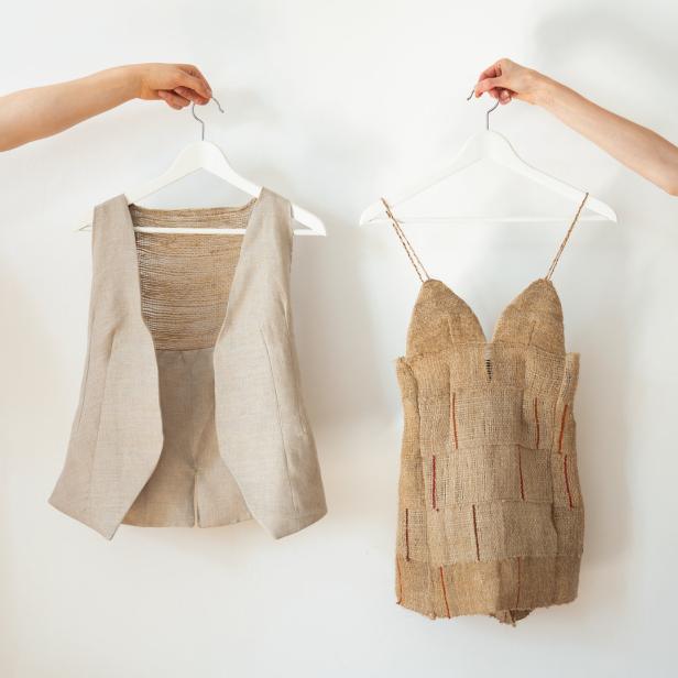 Zwei Hände halten eine Weste und ein Kleid aus grobem Stoff an Kleiderbügeln vor einer weißen Wand.